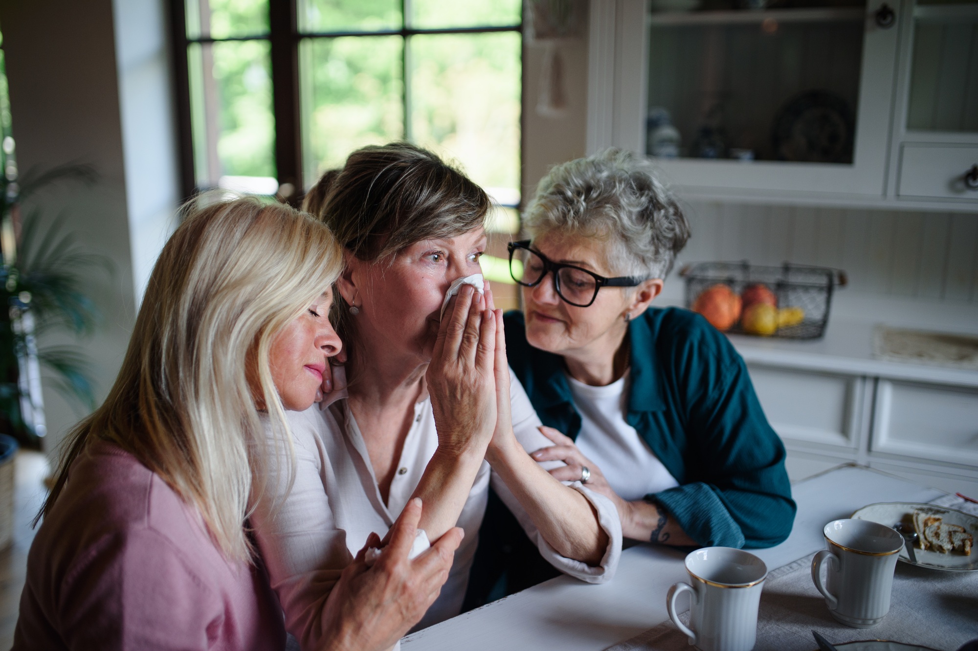Female senior friends consloing and supporting sad friend indoors at home.